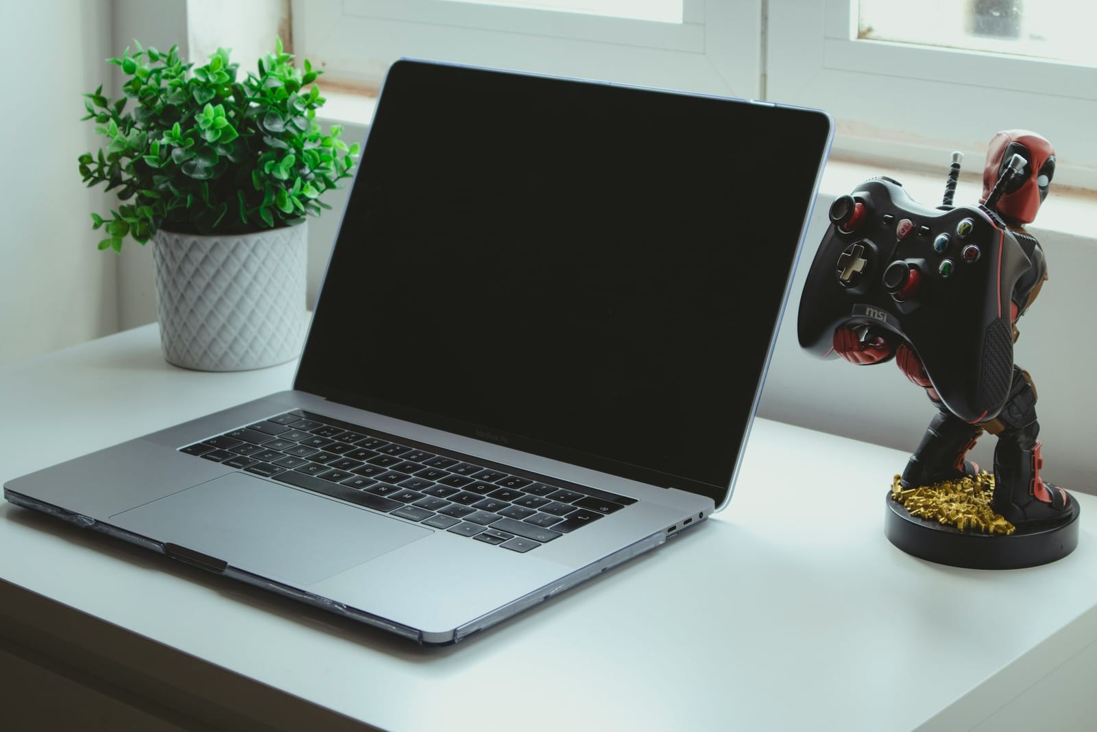 An image of a MacBook sitting on a white desk next to a plant and a video game controller on a stand that looks like the Marvel character Deadpool